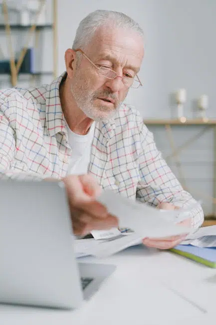 Elderly man reviewing documents at desk with computer.